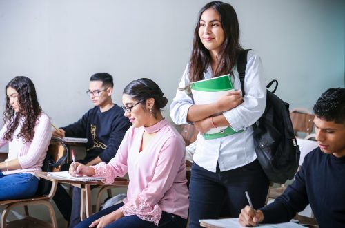A photograph of students sitting working in a room at individual desks. One student is standing up with a smile.