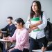 A photograph of students sitting working in a room at individual desks. One student is standing up with a smile.